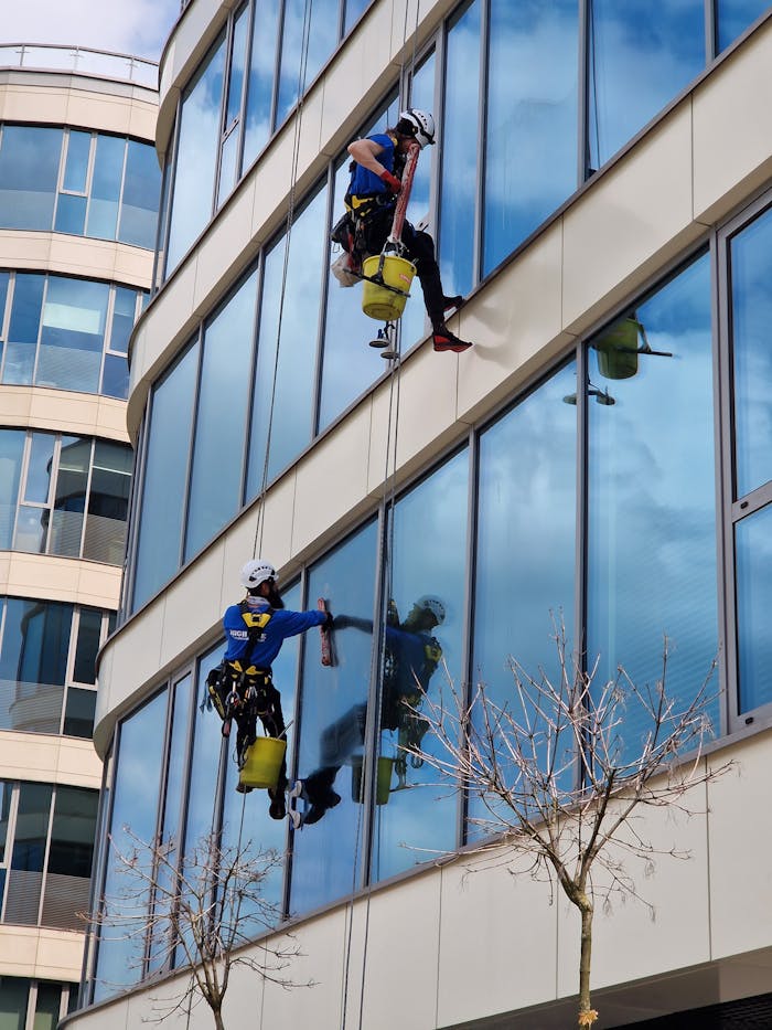 about-us-01 Two window cleaners using ropes on a modern skyscraper, showcasing urban professional maintenance.